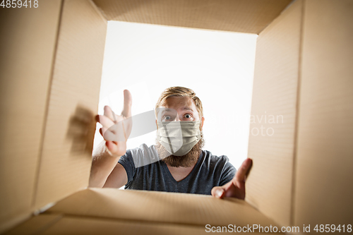 Image of Young man opening the huge postal package wearing protective face mask, contactless delivery