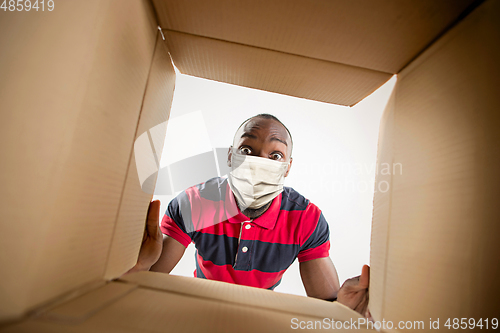 Image of Young man opening the huge postal package wearing protective face mask, contactless delivery