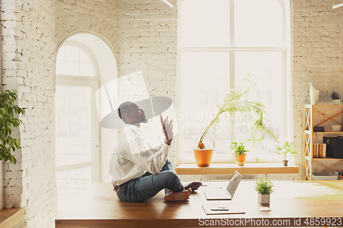 Image of Young african-american man doing yoga at home while being quarantine and freelance working