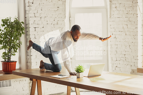 Image of Young african-american man doing yoga at home while being quarantine and freelance working