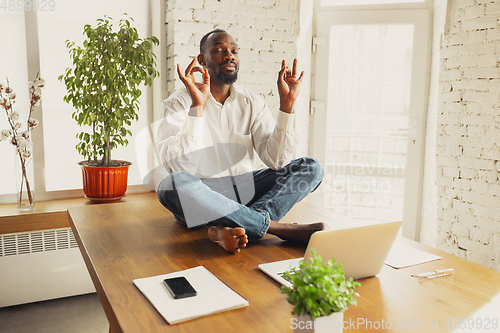 Image of Young african-american man doing yoga at home while being quarantine and freelance working