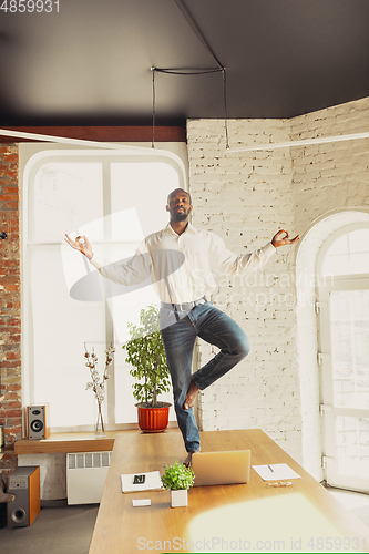 Image of Young african-american man doing yoga at home while being quarantine and freelance working