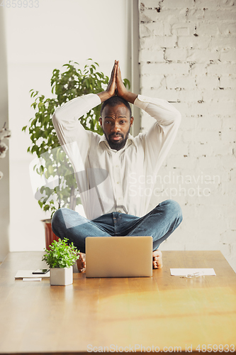 Image of Young african-american man doing yoga at home while being quarantine and freelance working