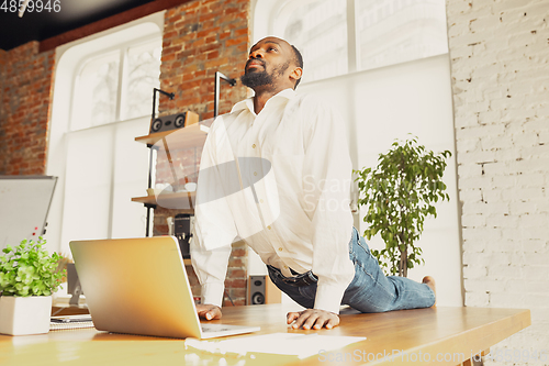 Image of Young african-american man doing yoga at home while being quarantine and freelance working