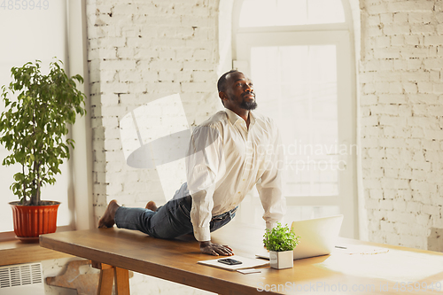 Image of Young african-american man doing yoga at home while being quarantine and freelance working