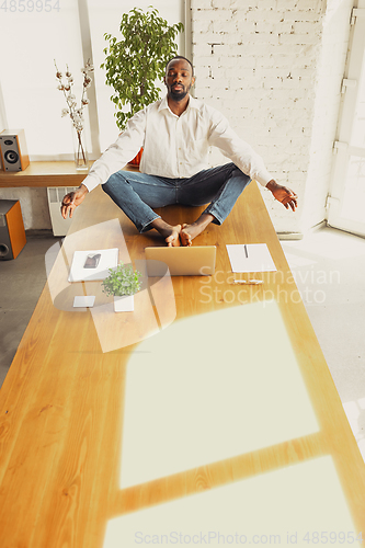 Image of Young african-american man doing yoga at home while being quarantine and freelance working