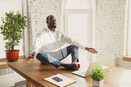 Image of Young african-american man doing yoga at home while being quarantine and freelance working