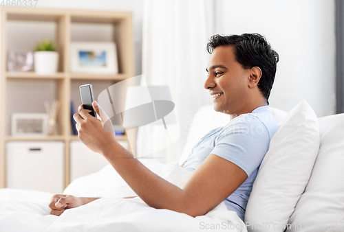 Image of happy indian man with smartphone in bed at home