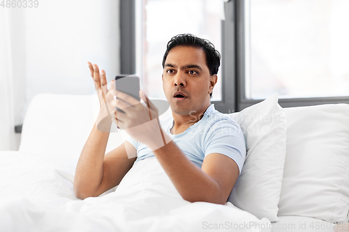 Image of angry indian man with smartphone in bed at home