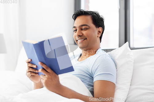 Image of happy indian man reading book in bed at home