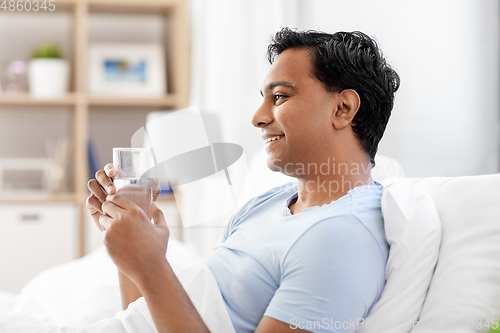 Image of happy indian man drinking water lying in bed