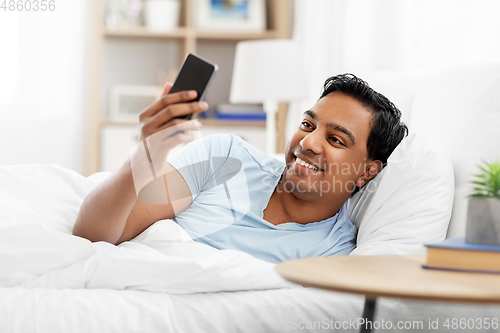 Image of happy indian man with smartphone in bed at home