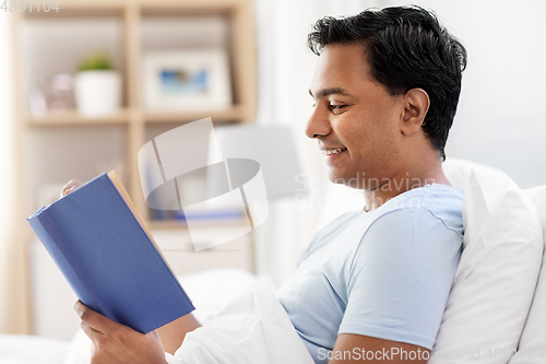 Image of happy indian man reading book in bed at home