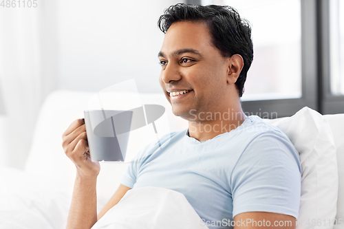 Image of happy indian man drinking coffee in bed at home