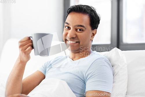 Image of happy indian man drinking coffee in bed at home