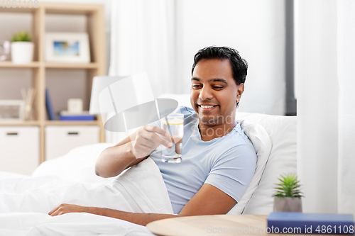 Image of happy indian man drinking water lying in bed