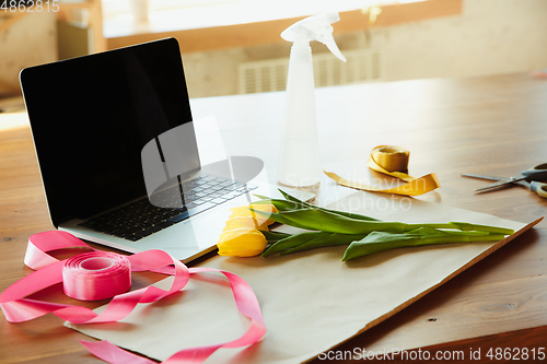 Image of Florist at work: table with tulips and daffodils, ribbons and coverage paper against laptop, prepared for workshop