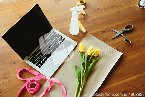 Image of Florist at work: table with tulips and daffodils, ribbons and coverage paper against laptop, prepared for workshop