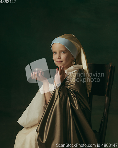 Image of Medieval little girl as a lady with a pearl earring on dark studio background. Concept of comparison of eras, childhood. Stylish creative design, art vision, new look of artwork.