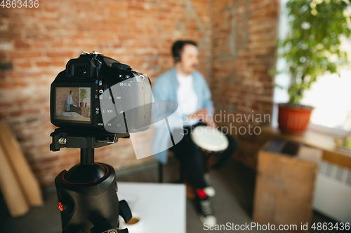 Image of Caucasian musician playing hand drum during online concert at home isolated and quarantined, focus on camera