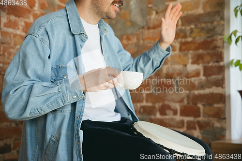 Image of Caucasian musician playing hand drum during online concert at home isolated and quarantined, greeting band and coffee drinking