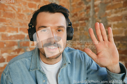 Image of Caucasian musician in headphones greeting audience before online concert at home isolated and quarantined, cheerful, smiling