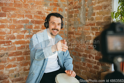 Image of Caucasian musician playing hand drum during online concert at home isolated and quarantined, smiling, talking to audience