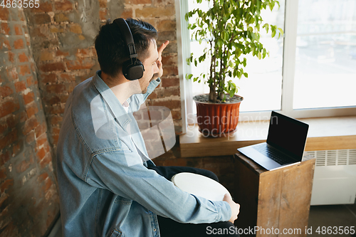 Image of Caucasian musician playing hand drum during online concert at home isolated and quarantined, greeting band or audience