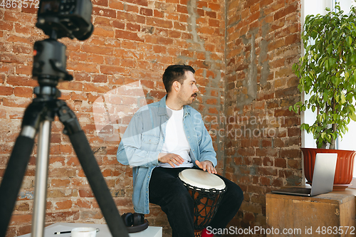Image of Caucasian musician playing hand drum during online concert at home isolated and quarantined, improvising