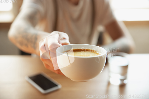 Image of Close up of male hands proposing cup of coffee, sitting at the table with smartphone