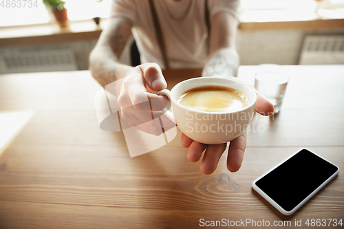 Image of Close up of male hands proposing cup of coffee, sitting at the table with smartphone