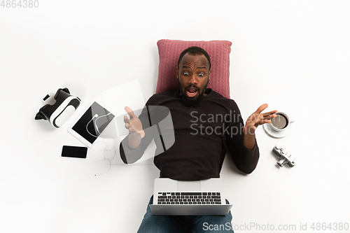 Image of Emotional african-american man using laptop surrounded by gadgets isolated on white studio background, technologies. Shocked