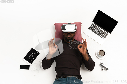 Image of Emotional african-american man using VR-headset surrounded by gadgets isolated on white studio background, technologies. Nice, happy