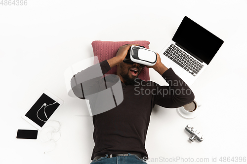 Image of Emotional african-american man using VR-headset surrounded by gadgets isolated on white studio background, technologies. Emotional playing