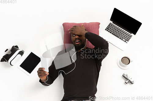 Image of Emotional african-american man using phone surrounded by gadgets isolated on white studio background, technologies. Shocked