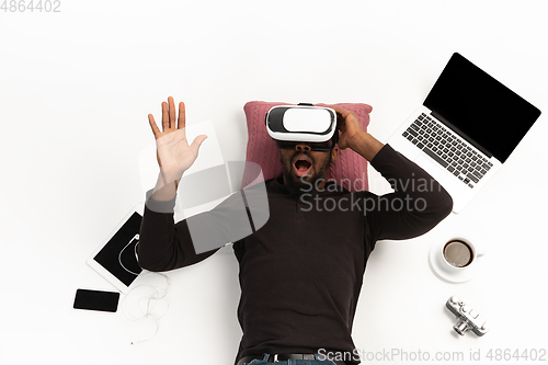 Image of Emotional african-american man using VR-headset surrounded by gadgets isolated on white studio background, technologies. Emotional playing