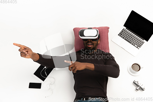 Image of Emotional african-american man using VR-headset surrounded by gadgets isolated on white studio background, technologies. Emotional playing