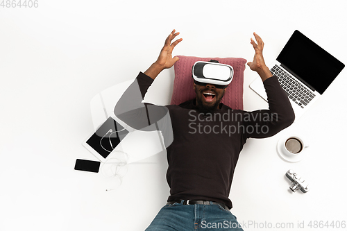 Image of Emotional african-american man using VR-headset surrounded by gadgets isolated on white studio background, technologies. Emotional playing