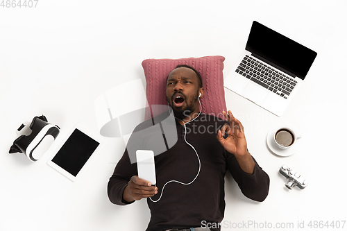 Image of Emotional african-american man using phone surrounded by gadgets isolated on white studio background, technologies connecting people. Inspired singing