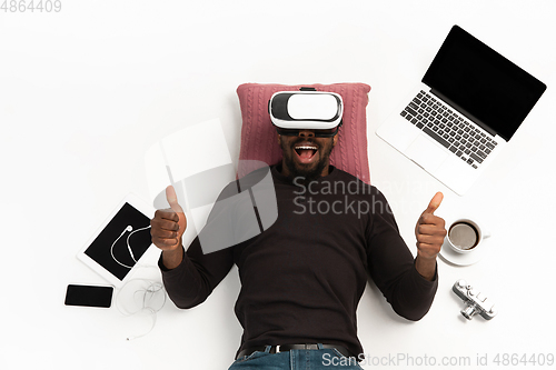Image of Emotional african-american man using VR-headset surrounded by gadgets isolated on white studio background, technologies. Thumbs up, happy