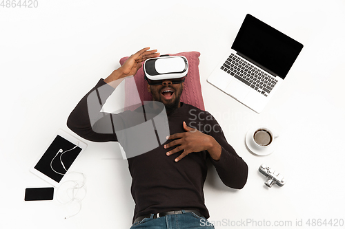 Image of Emotional african-american man using VR-headset surrounded by gadgets isolated on white studio background, technologies. Emotional playing