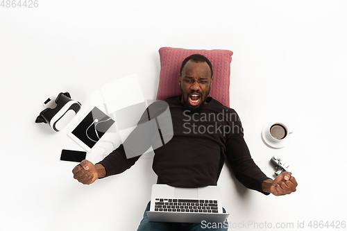 Image of Emotional african-american man using laptop surrounded by gadgets isolated on white studio background, technologies. Winning