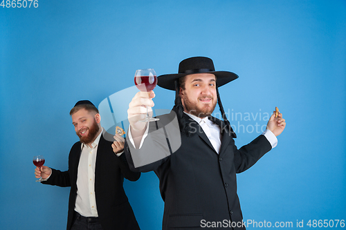 Image of Portrait of a young orthodox jewish men isolated on blue studio background, meeting the Passover, eating Aman\'s ears with wine