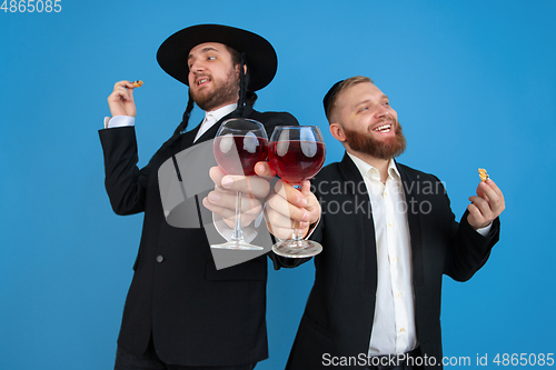 Image of Portrait of a young orthodox jewish men isolated on blue studio background, meeting the Passover, eating Aman\'s ears with wine