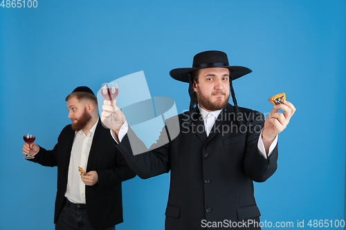 Image of Portrait of a young orthodox jewish men isolated on blue studio background, meeting the Passover, eating Aman\'s ears with wine