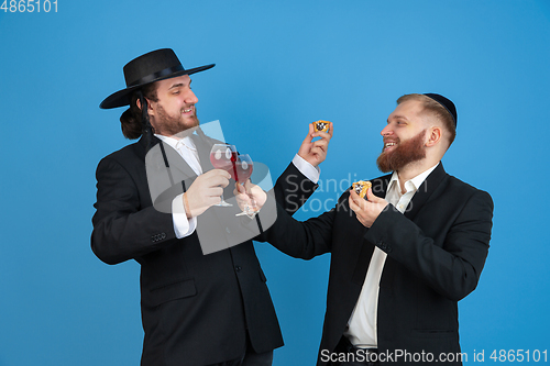 Image of Portrait of a young orthodox jewish men isolated on blue studio background, meeting the Passover, eating Aman\'s ears with wine