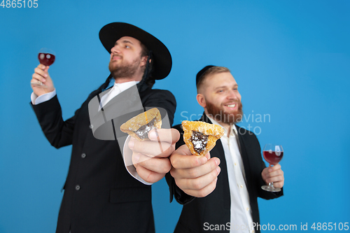 Image of Portrait of a young orthodox jewish men isolated on blue studio background, meeting the Passover, eating Aman\'s ears with wine