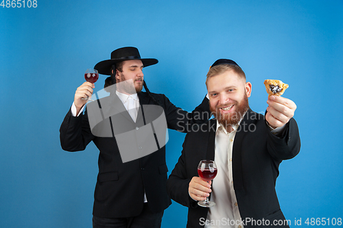 Image of Portrait of a young orthodox jewish men isolated on blue studio background, meeting the Passover, eating Aman\'s ears with wine