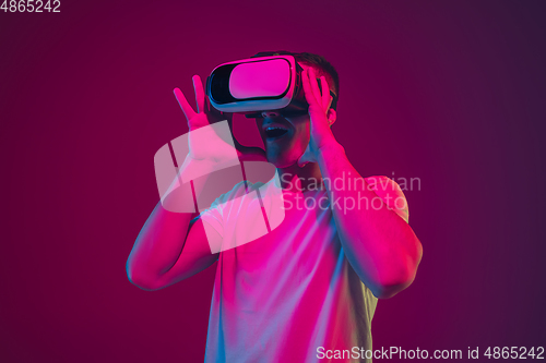 Image of Caucasian man\'s portrait isolated on pink-purple studio background in neon light, playing with VR-headset