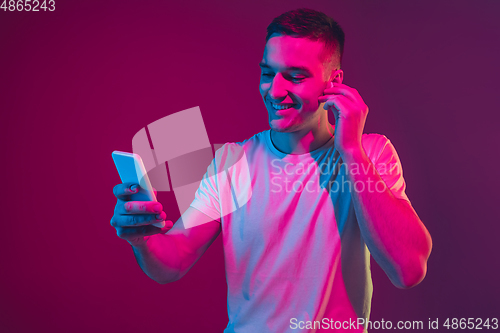 Image of Caucasian man\'s portrait isolated on pink-purple studio background in neon light, with smartphone and earphones